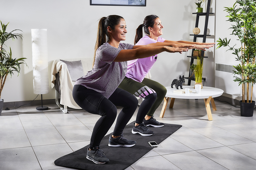 2 femmes dans leur salon en train de réaliser un squat avec le sourire sur un tapis de fitness en suivant l'application FizzUp sur leur téléphone
